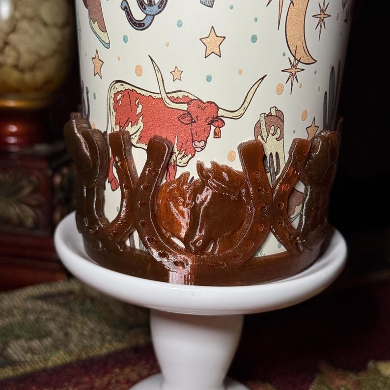 K&ouml;nnte beinhalten: Ein brauner Becherhalter mit einem Hufeisen- und Pferdedesign. Der Becher hat einen cremefarbenen Hintergrund mit einem Cartoon-Longhorn, Sternen und anderen Illustrationen zum Thema Western. Der Halter steht auf einem wei&szlig;en Sockel.