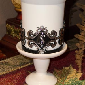 May include: White ceramic candle holder with a black band and a silver decorative trim featuring a stylized letter "S".