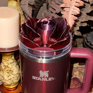 May include: A burgundy stainless steel travel mug with a rose-shaped lid. The mug has a silver band around the middle and the Stanley logo is engraved on the front.