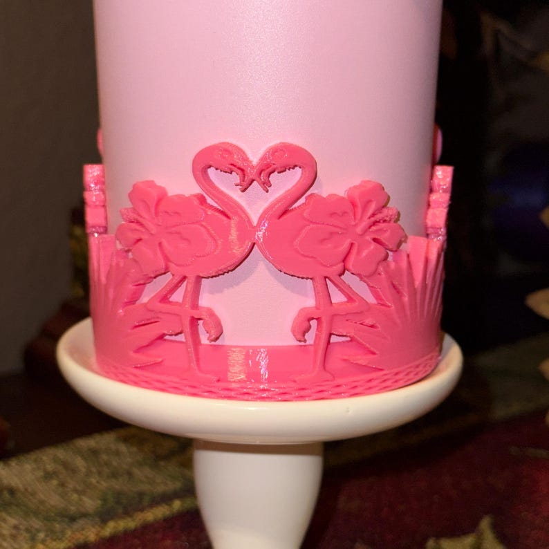 May include: A light pink cylindrical container featuring a raised, bright pink decorative wrap. The wrap displays two flamingos facing each other with their necks forming a heart shape, flanked by hibiscus flowers and tropical foliage patterns.