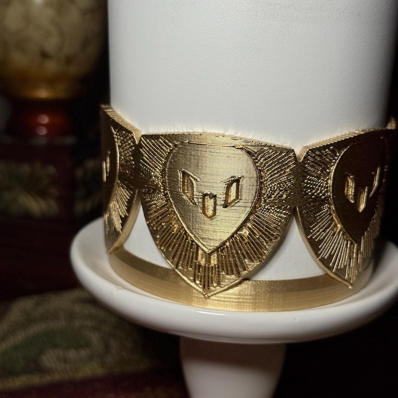 May include: A white candle holder with a gold-colored decorative band. The band features a repeating heart-shaped design with a sunburst pattern. The candle holder sits on a white pedestal.