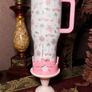 May include: A white HydroJug tumbler with a pink handle and lid, featuring a repeating pattern of pink flamingos, palm trees, vintage cars, suitcases, and beach umbrellas. The base is held by a pink stand decorated with two facing flamingos.