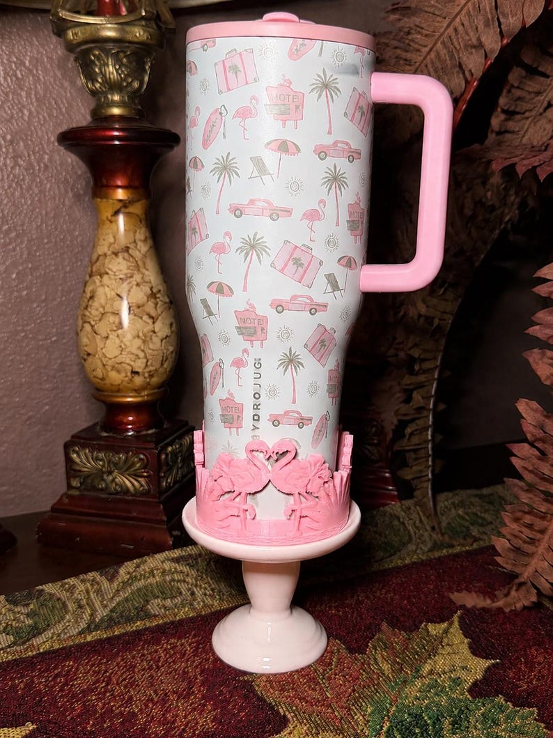May include: A white HydroJug tumbler with a pink handle and lid, featuring a repeating pattern of pink flamingos, palm trees, vintage cars, suitcases, and beach umbrellas. The base is held by a pink stand decorated with two facing flamingos.