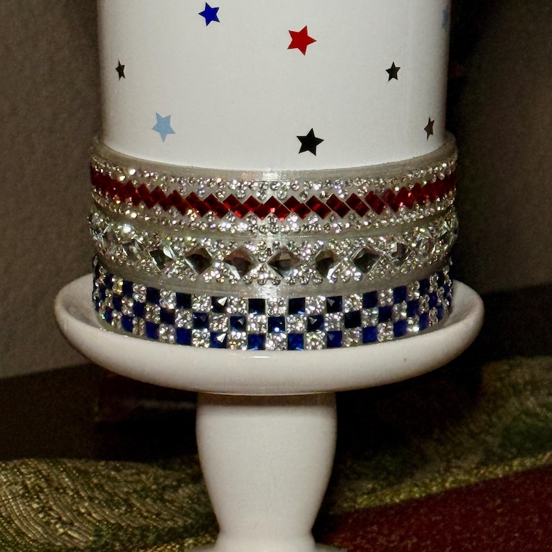 May include: A white pedestal candle holder with a decorative design. The candle holder features a white base and stem, with a cylindrical top adorned with red, silver, and blue jewel-like embellishments. The top has a pattern of red, blue, and black stars.