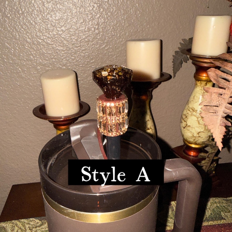 May include: A brown travel mug with a decorative straw topper. The topper has a faceted, amber-colored gem design with gold flecks and a clear, crystal-like base. The text "Style A" is displayed on the mug.