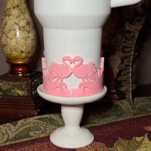 May include: A white pedestal mug featuring a pink wrap-around band with a raised relief design of two flamingos facing each other, their necks forming a heart shape, with additional palm leaf motifs integrated into the pink decorative band.