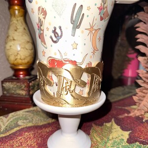 K&ouml;nnte beinhalten: Ein wei&szlig;er Becher mit einem goldfarbenen dekorativen Halter. Der Becher hat ein Western-Design mit Stiefeln, Kakteen und Sternen. Der Halter ist mit einem sich wiederholenden Stiefelmuster verziert. Der Becher steht auf einem wei&szlig;en Sockel.