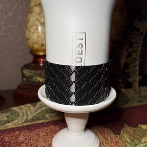 May include: A white travel mug with the word "DESTI" printed vertically. The mug sits on a white pedestal with a black patterned sleeve. The mug has a handle and a black lid.