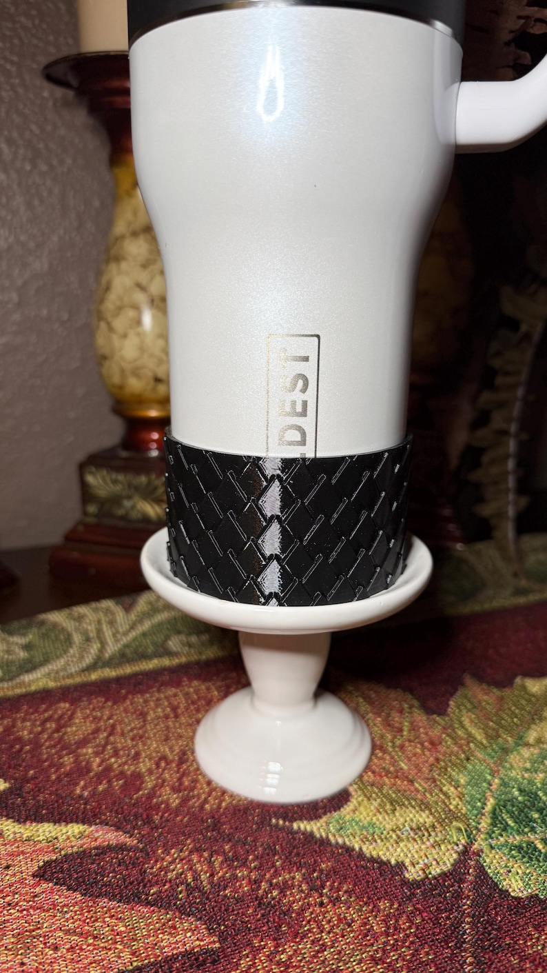 May include: A white travel mug with the word "DESTI" printed vertically. The mug sits on a white pedestal with a black patterned sleeve. The mug has a handle and a black lid.