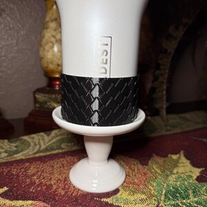May include: A white ceramic candle holder with a black patterned band around the middle. The candle holder has a pedestal base and the word "DEST" is printed on the side.