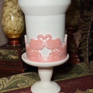 May include: A white pedestal cup featuring a wrap-around relief design of two pink flamingos facing each other with their necks forming a heart shape. The base of the cup is flared, and the flamingo motif includes palm fronds and sun rays.