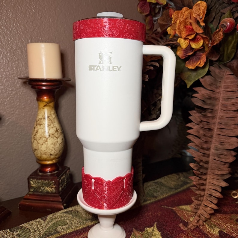 May include: A white Stanley tumbler featuring a handle and a red lid with a paisley pattern. The tumbler includes a matching red decorative base accessory with a detailed embossed design, positioned on a white pedestal stand for display purposes.