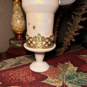May include: A white candle holder with a gold crown-shaped accent. The candle holder has a pearlescent finish with starburst designs. The base is white and round, and the holder is on a decorative surface.