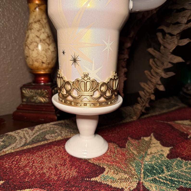 May include: A white candle holder with a gold crown-shaped accent. The candle holder has a pearlescent finish with starburst designs. The base is white and round, and the holder is on a decorative surface.