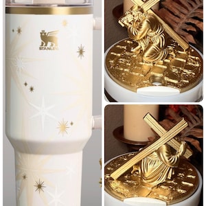 Puede incluir: Un vaso Stanley blanco con detalles dorados y dise&ntilde;os de estrellas. El vaso tiene una tapa transparente y un asa. Una figura religiosa dorada representa a una persona arrodillada que lleva una cruz.