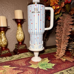 May include: A white Stanley tumbler with a blue and white striped pattern and red star accents. The tumbler has a white handle and is displayed on a white pedestal with a star design. The Stanley logo is visible.