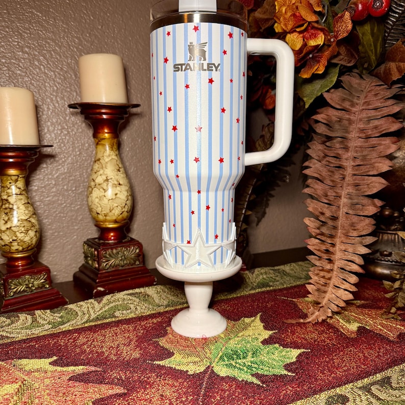 May include: A white Stanley tumbler with a blue and white striped pattern and red star accents. The tumbler has a white handle and is displayed on a white pedestal with a star design. The Stanley logo is visible.
