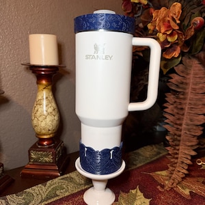 May include: A white Stanley tumbler with a handle, featuring a dark blue paisley-patterned lid and a matching dark blue decorative base accessory. The tumbler has the Stanley logo printed in silver on the front of the white cylindrical body.