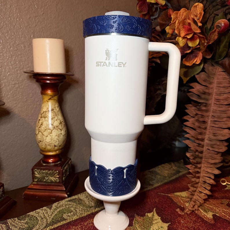 May include: A white Stanley tumbler with a handle, featuring a dark blue paisley-patterned lid and a matching dark blue decorative base accessory. The tumbler has the Stanley logo printed in silver on the front of the white cylindrical body.