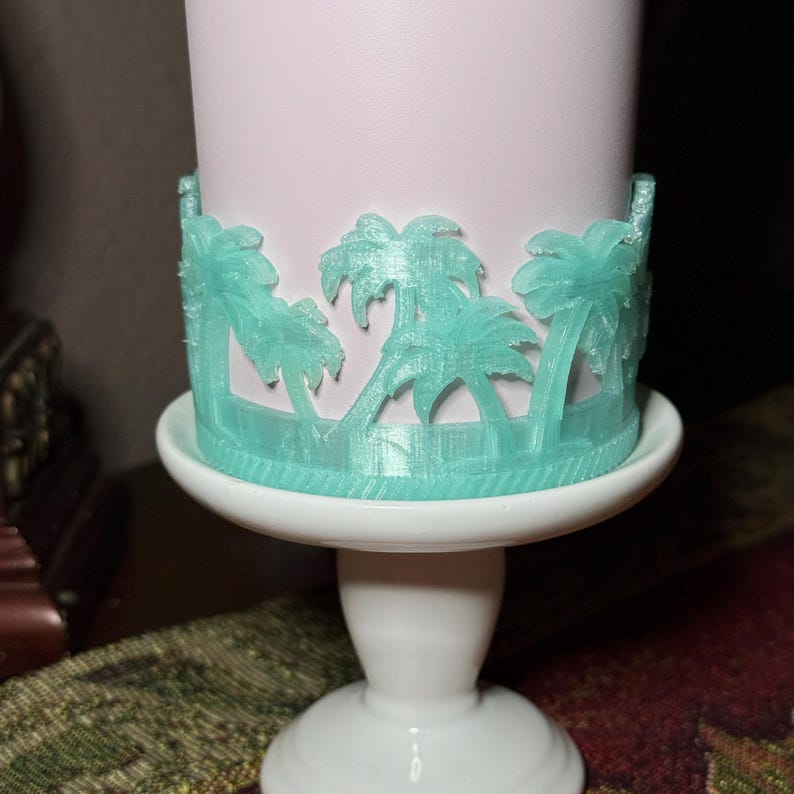 May include: A light pink cylindrical container featuring a wrap-around 3D-printed mint green palm tree design. The container is positioned on a small white pedestal stand with a smooth, rounded base and a central column support structure.