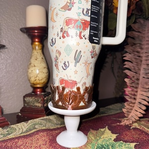 K&ouml;nnte beinhalten: Ein 1,13-Liter-Becher mit Western-Design, mit Stiefeln, Kakteen und Langhorn-Rindern. Er steht auf einem wei&szlig;en Sockel mit einem braunen dekorativen Halter.