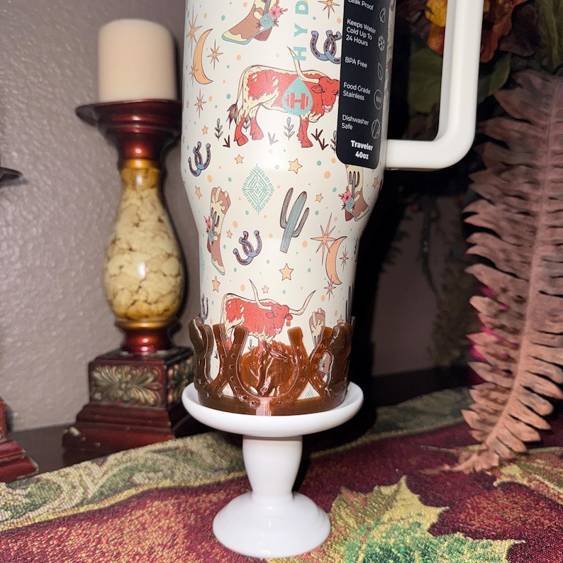 K&ouml;nnte beinhalten: Ein 1,13-Liter-Becher mit Western-Design, mit Stiefeln, Kakteen und Langhorn-Rindern. Er steht auf einem wei&szlig;en Sockel mit einem braunen dekorativen Halter.