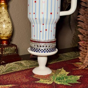 May include: A white tumbler with blue stripes and red star accents, paired with a stack of decorative bracelets on a small white pedestal. The bracelets feature red, silver, and blue designs. The tumbler has a white handle.