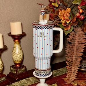 May include: A white Stanley tumbler with blue vertical stripes and red star accents. The tumbler has a white handle and is decorated with a jeweled rim and base. A decorative stopper with a crystal top is inserted into the tumbler.
