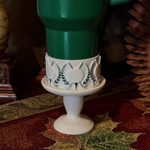 May include: A white ceramic candle holder with a green candle. The candle holder has a decorative band with white medallions and green leaf accents. The base is round and the candle holder is on a patterned surface.