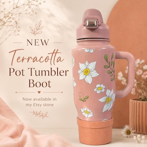 May include: A pink tumbler with a floral pattern featuring white flowers and green leaves, topped with a matching pink lid and handle. The base is fitted with a terracotta-colored silicone boot. Text on the left reads: NEW Terracotta Pot Tumbler Boot.