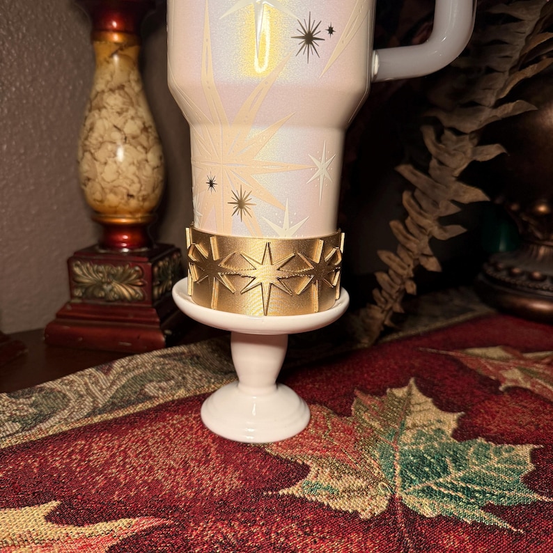 May include: A white travel mug with a gold star design and a gold band. The mug is resting on a small white pedestal. The background includes a decorative table runner with a leaf pattern.