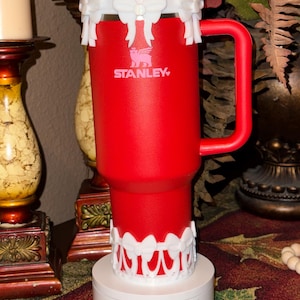 May include: Red Stanley travel mug with a white bow and lace trim around the lid and base. The mug is on a white pedestal with the words "Stone Oak Creatives" printed on the front.