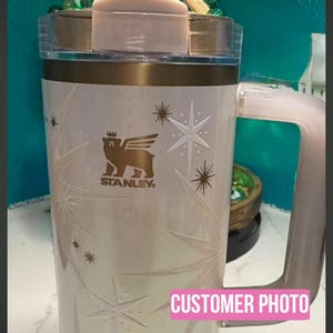Puede incluir: Un vaso Stanley blanco con tapa y asa doradas, con una base decorativa de madera. El vaso tiene un logotipo y dise&ntilde;os de estrellas. Una estatua dorada de una persona que lleva una cruz est&aacute; encima. El texto "CUSTOMER PHOTO" es visible.