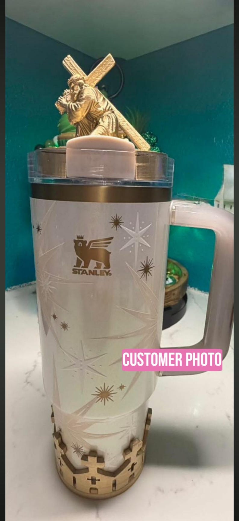 Puede incluir: Un vaso Stanley blanco con tapa y asa doradas, con una base decorativa de madera. El vaso tiene un logotipo y dise&ntilde;os de estrellas. Una estatua dorada de una persona que lleva una cruz est&aacute; encima. El texto "CUSTOMER PHOTO" es visible.