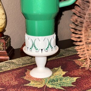 May include: A green glittery golf-themed mug on a white pedestal. The mug has a white band with golf ball and club designs. The pedestal is white and has a round base. The mug is on a table runner with a leaf pattern.