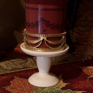 May include: A burgundy-colored candle holder with a gold-colored decorative band. The candle holder sits on a white pedestal base. The candle holder has a decorative design around the top.