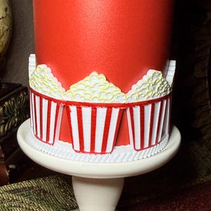 Pu&ograve; includere: Un oggetto cilindrico rosso decorato con un bordo ripetitivo di contenitori per popcorn tridimensionali a strisce bianche e rosse. Ogni contenitore presenta una parte superiore di popcorn bianchi testurizzati con accenti gialli alla base.