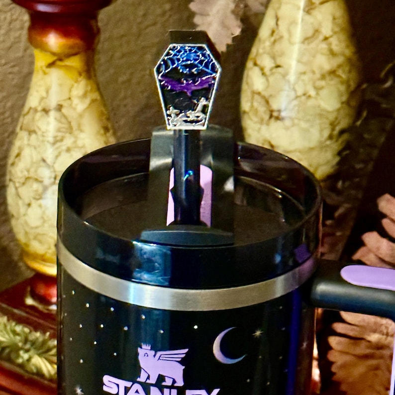 May include: Black Stanley tumbler with a decorative lid featuring a colorful, abstract design. The tumbler has a silver band and a handle. The Stanley logo and a crescent moon are visible on the tumbler.