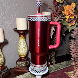 May include: A red Stanley + Starbucks travel mug with a pink handle and a decorative rim. The mug is embellished with rhinestones and sits on a white stand. A decorative stem with a crystal topper is also present.