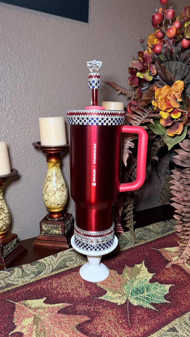 May include: A red Stanley + Starbucks travel mug with a pink handle and a decorative rim. The mug is embellished with rhinestones and sits on a white stand. A decorative stem with a crystal topper is also present.