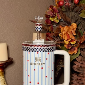 May include: A Stanley tumbler with a white handle, adorned with red stars and blue stripes. The rim is embellished with red, white, and blue rhinestones. A decorative crystal topper sits on the lid. The Stanley logo is visible.