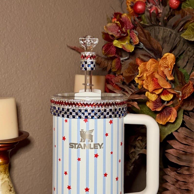 May include: A Stanley tumbler with a white handle, adorned with red stars and blue stripes. The rim is embellished with red, white, and blue rhinestones. A decorative crystal topper sits on the lid. The Stanley logo is visible.