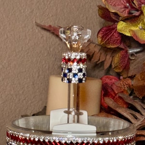 May include: A decorative bottle stopper with a clear, faceted crystal top and a cylindrical body adorned with red, white, and blue rhinestones. The stopper is designed to fit into a bottle, adding a touch of elegance.