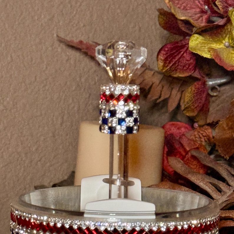 May include: A decorative bottle stopper with a clear, faceted crystal top and a cylindrical body adorned with red, white, and blue rhinestones. The stopper is designed to fit into a bottle, adding a touch of elegance.