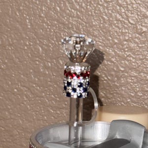 May include: A decorative bottle stopper with a clear, faceted crystal top. The stopper features a band of red, white, and blue rhinestones. The stopper is attached to a clear, thin rod.