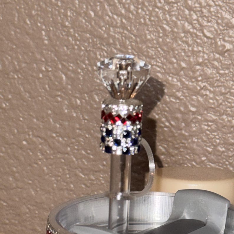 May include: A decorative bottle stopper with a clear, faceted crystal top. The stopper features a band of red, white, and blue rhinestones. The stopper is attached to a clear, thin rod.