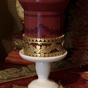 May include: A burgundy candle holder with a gold filigree heart design. The candle holder sits on a white pedestal base. The candle holder has a decorative pattern around the top.