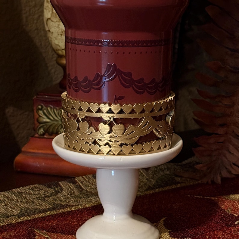 May include: A burgundy candle holder with a gold filigree heart design. The candle holder sits on a white pedestal base. The candle holder has a decorative pattern around the top.