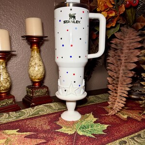 May include: White Stanley tumbler with a handle, decorated with red, blue, and black stars. The tumbler has the Stanley logo and is placed on a white star-shaped base. A decorative item for home or office.