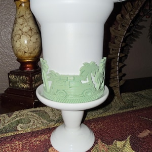May include: A white pedestal cup featuring a light green wrap-around relief design. The raised decoration depicts a vehicle with a surfboard on its roof, surrounded by palm trees. The cup has a smooth finish and a flared base, standing on a stem.